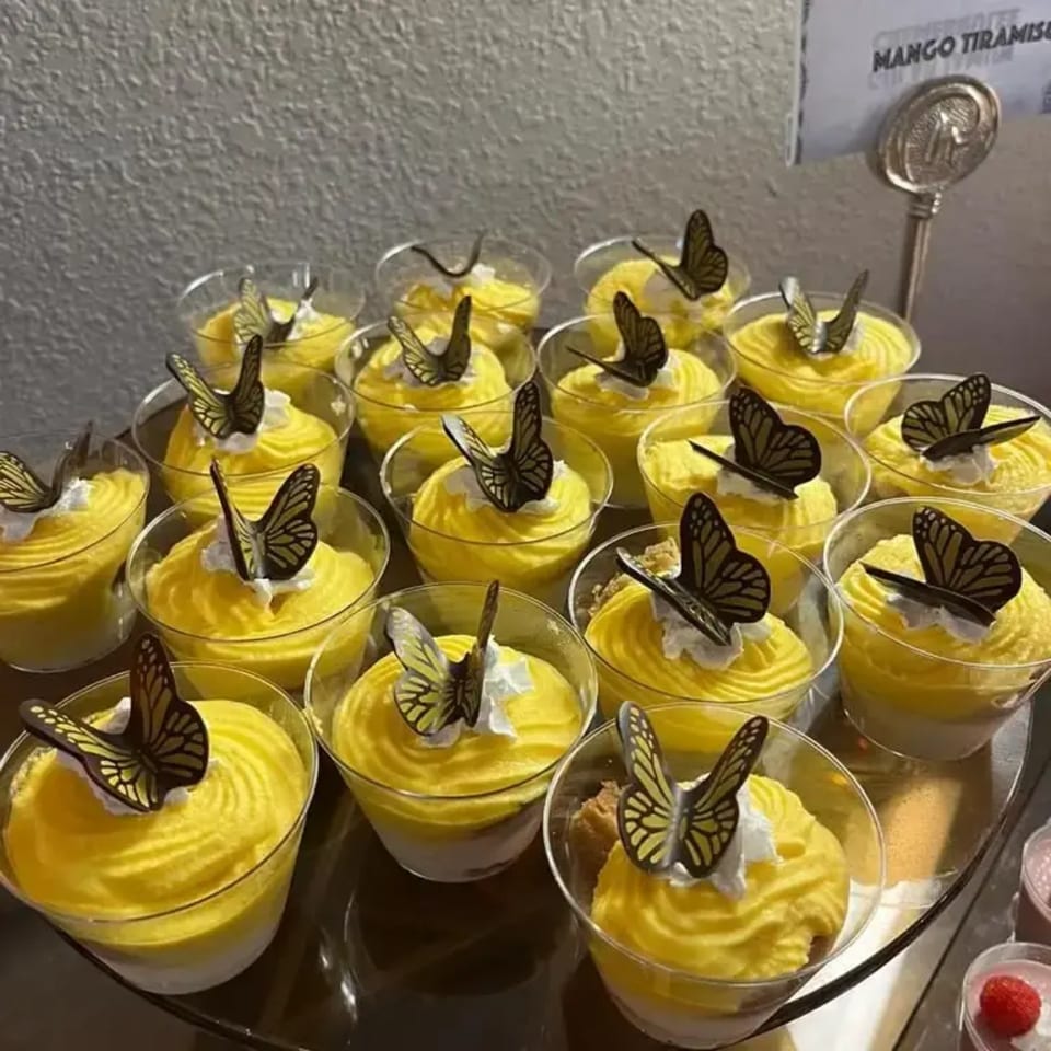 Numerous mango tiramisu desserts in clear cups, topped with whipped cream and chocolate butterflies.