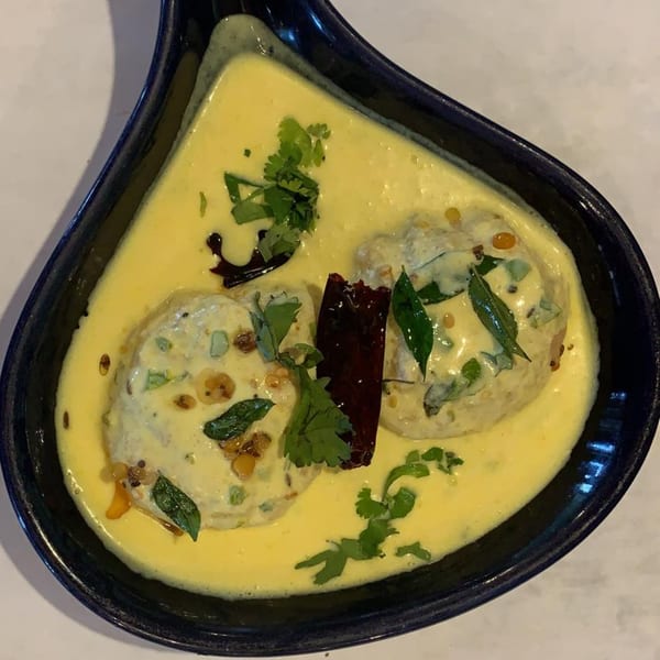 Two Indian dumplings in a creamy yellow sauce, garnished with herbs and dried chilies in a black dish.