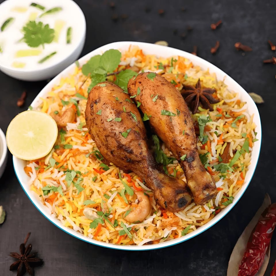 A vibrant bowl of Indian chicken biryani with two drumsticks, rice, cashews, and fresh herbs.