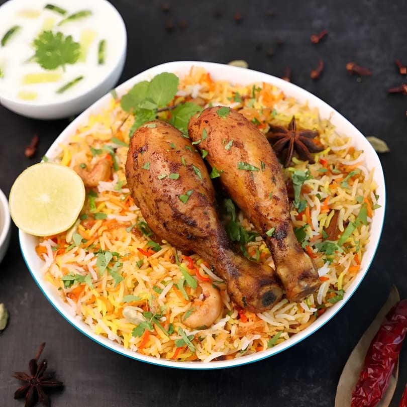 Chicken Biryani with two drumsticks, rice, cashews, and herbs, served with raita and lemon.